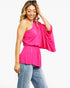 A woman modeling a bright pink one-shoulder top with a gathered waist and wide draped sleeves, paired with blue jeans, against a plain white background.