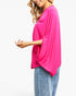 A woman wearing a bright pink, loose-fitting short-sleeve top with wide draped sleeves and light blue jeans, shown from a side profile against a plain white background.
