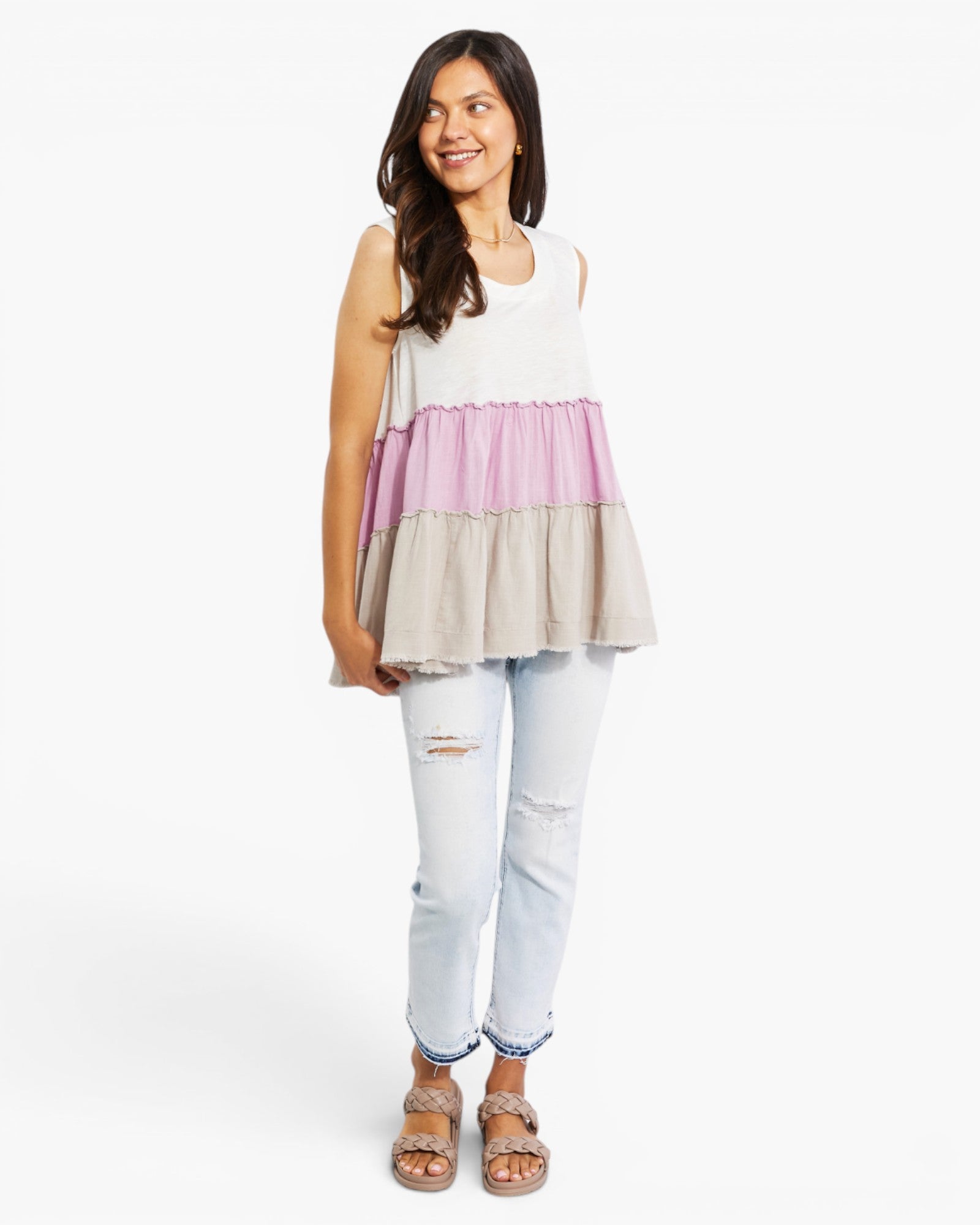 A woman modeling a sleeveless tiered tunic top with horizontal stripes in white, dusty pink, and light beige, paired with light-wash distressed jeans and brown sandals, against a plain white studio background.