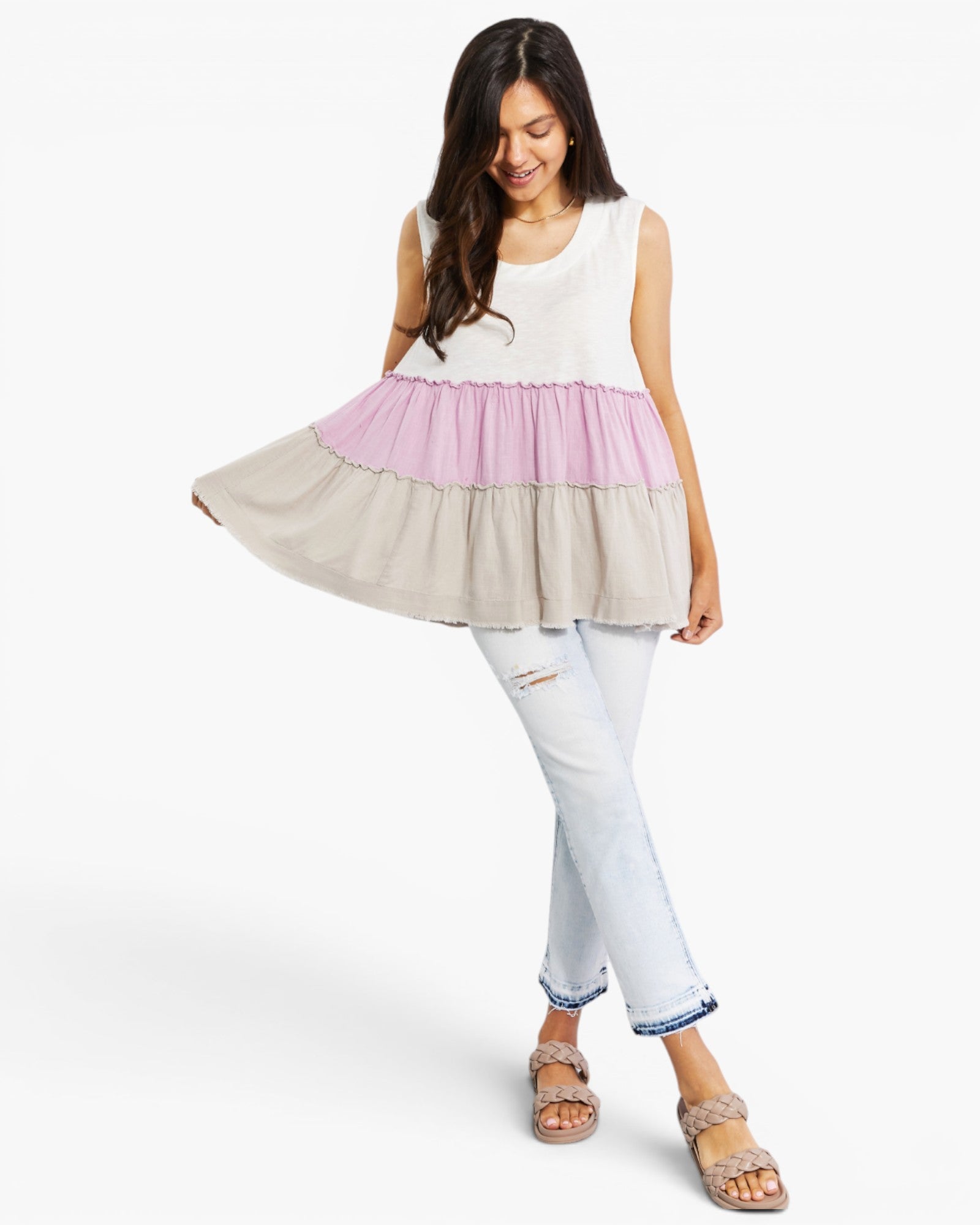 A woman modeling a sleeveless tiered tunic top with a white upper section, a light pink middle band, and a light beige flared lower skirt, paired with light-wash jeans and sandals on a white studio background.
