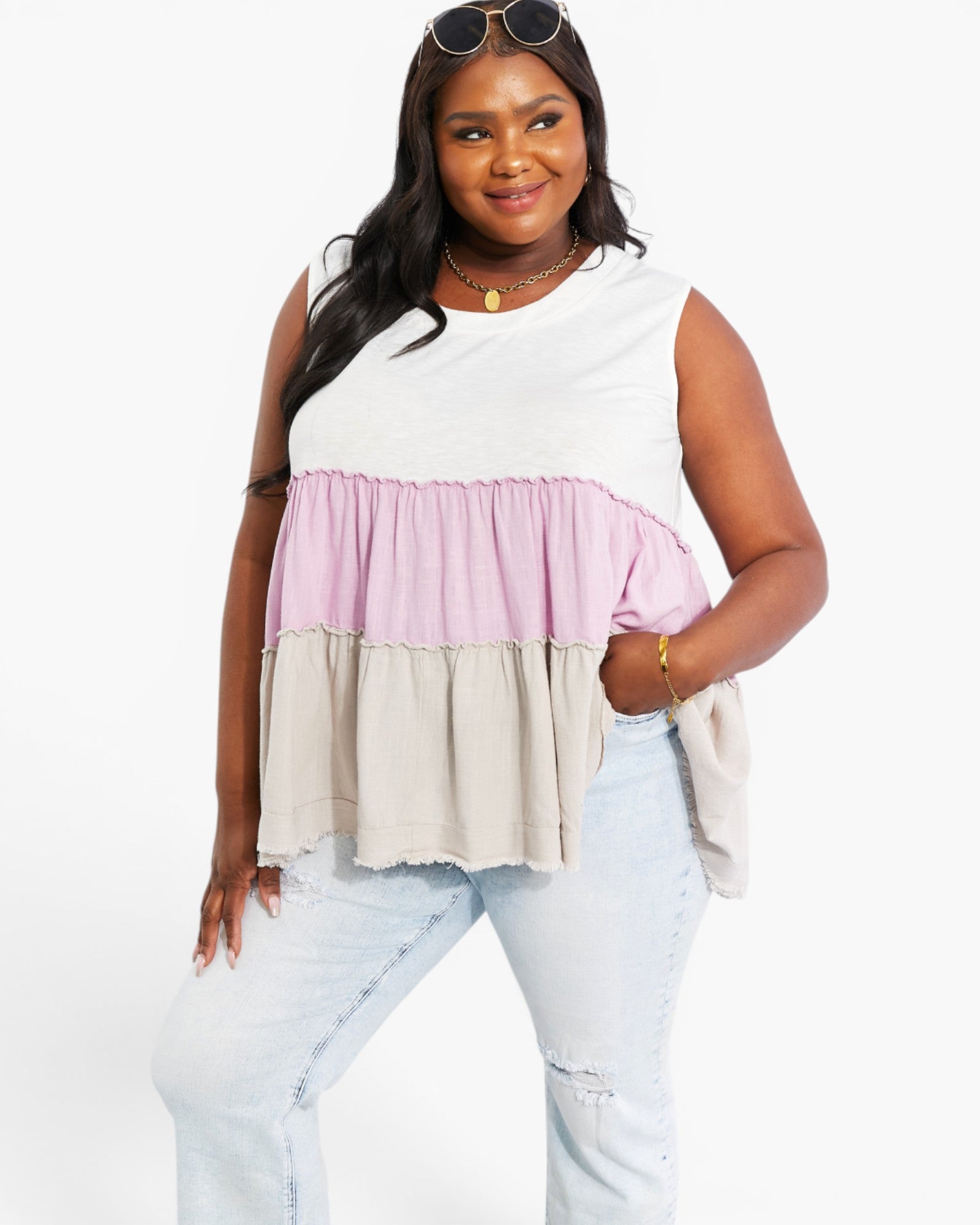 A woman modeling a sleeveless tiered top with a white upper section, a light pink ruffled band, and a light beige lower ruffle, paired with light-wash distressed jeans against a plain white background.