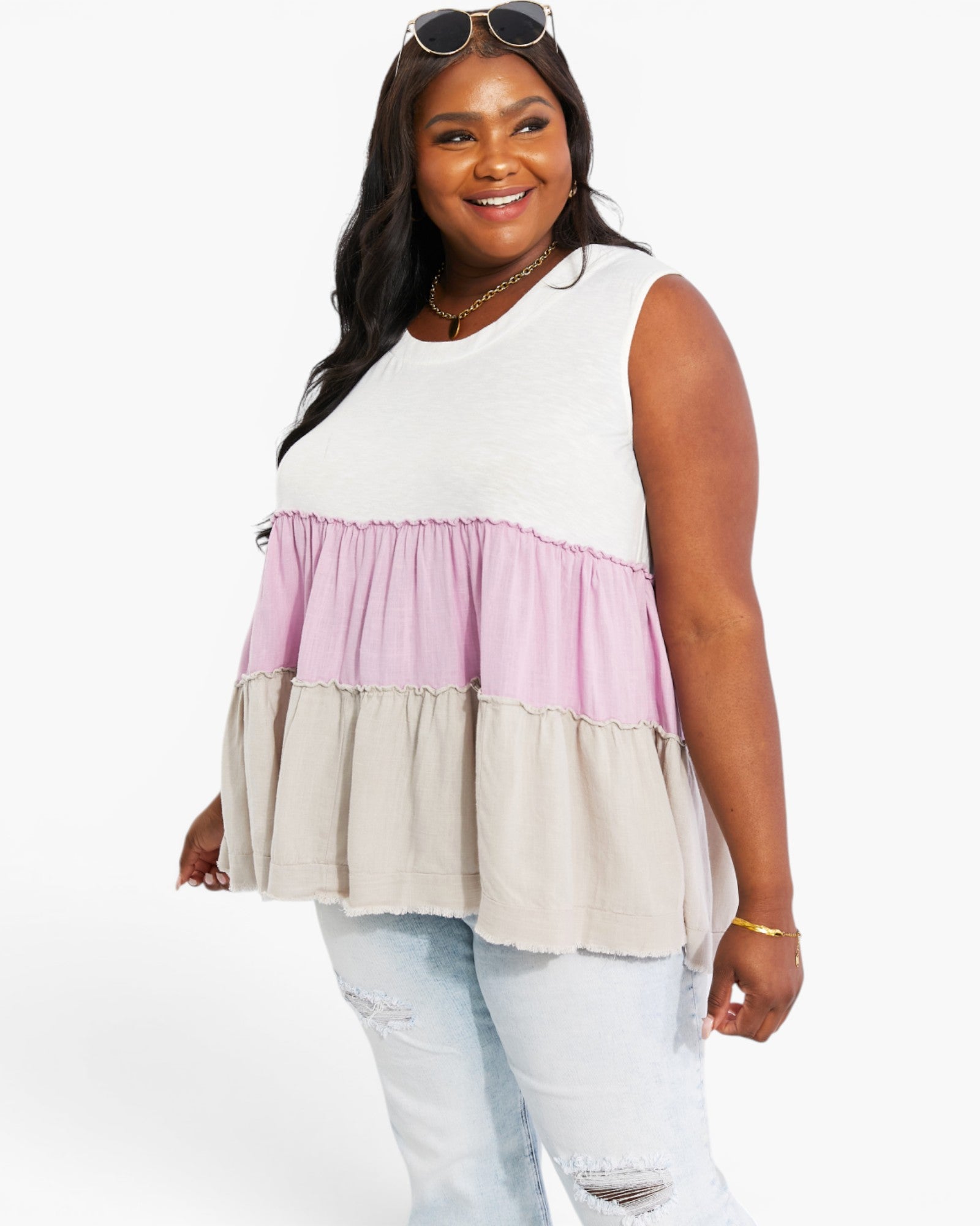 A woman modeling a sleeveless tiered peplum top with horizontal color-blocked ruffles in white, dusty pink, and light beige, paired with light-wash distressed jeans against a plain white studio background.
