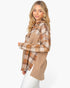 A woman modeling a long-sleeve button-up shirt with a mixed design featuring a brown and beige plaid top half and a solid tan textured bottom half, paired with white pants against a light gray studio background.
