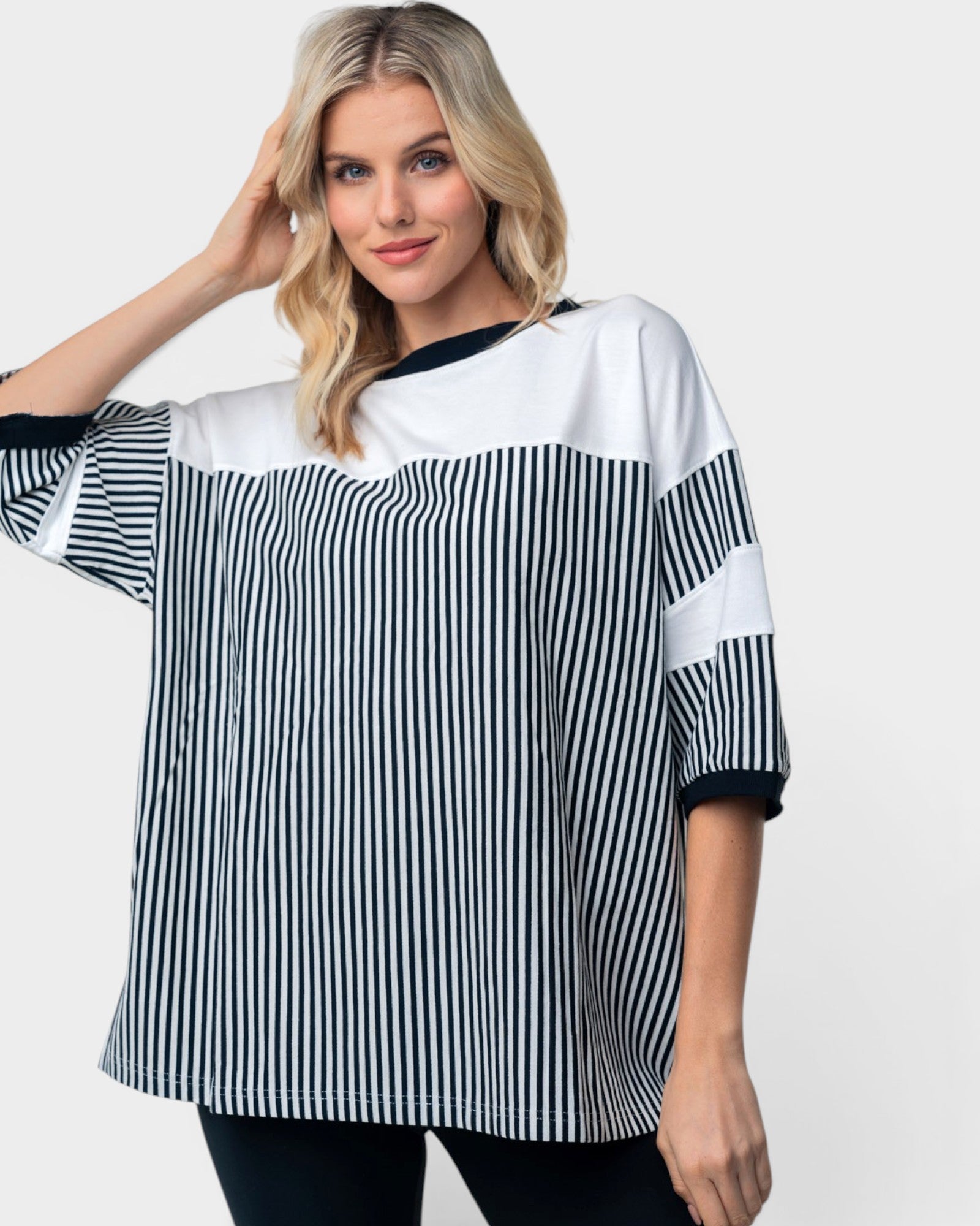 A woman modeling a loose, oversized short-sleeve top with a color-blocked design featuring vertical black and white stripes on the lower half, solid white on the upper chest and shoulders, and solid black trim at the neckline and sleeve cuffs.