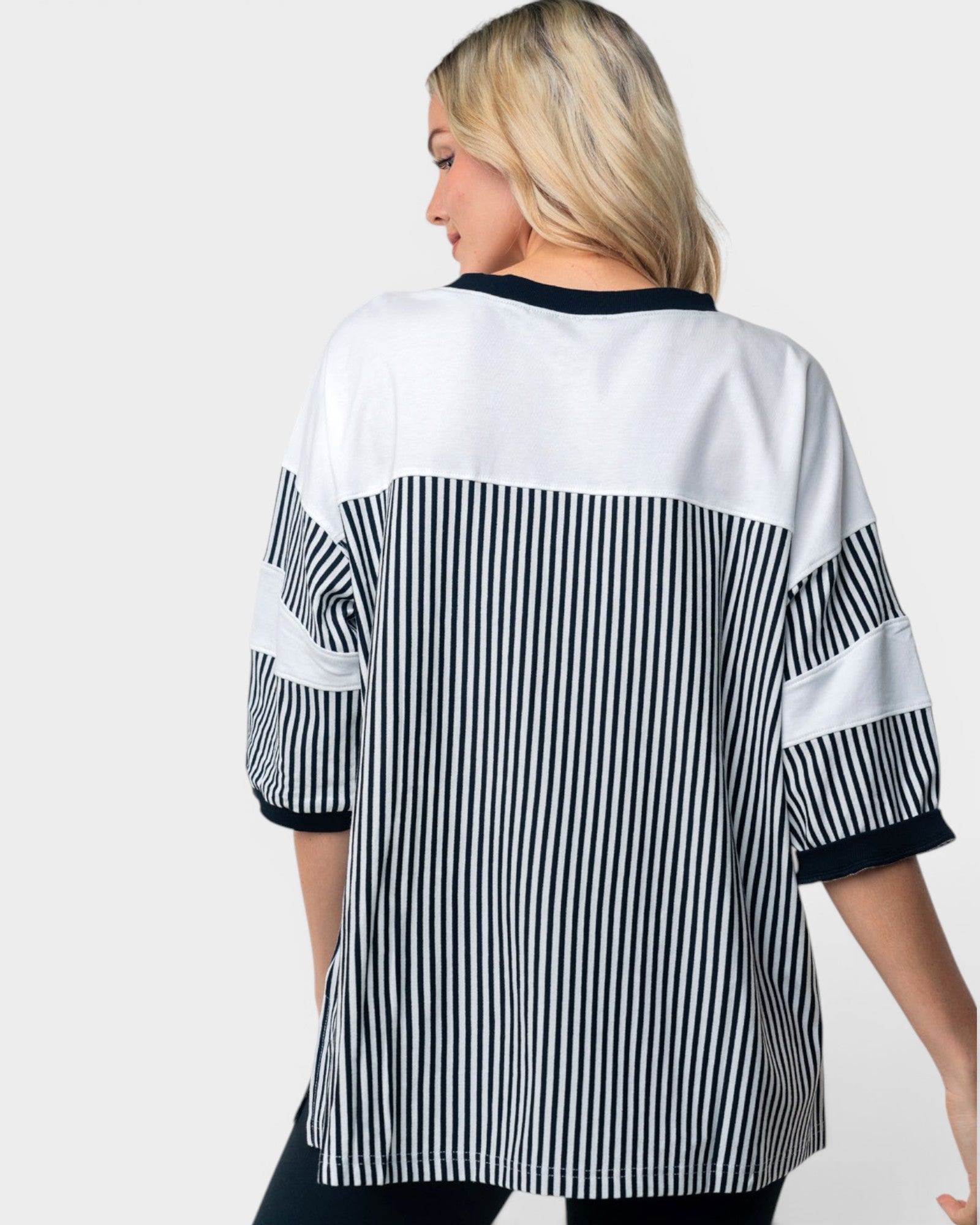 A woman shown from the back wearing an oversized short-sleeve top with a color-blocked design: white upper back and shoulders, black-and-white vertical stripes across the center and sleeves, and black trim at the neckline and sleeve cuffs, against a light gray studio background.