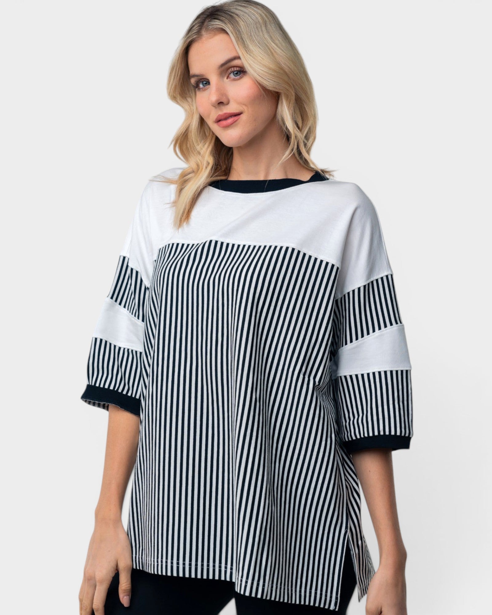 A woman wearing a color-blocked knit top with vertical black and white stripes on the lower half and sleeves, solid white on the upper half, and solid black trim on the crew neck and cuffs, against a light gray background.
