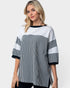 A woman wearing a color-blocked knit top with vertical black and white stripes on the lower half and sleeves, solid white on the upper half, and solid black trim on the crew neck and cuffs, against a light gray background.
