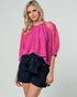 Woman wearing a pink blouse with sheer sleeves and black shorts on a light gray background