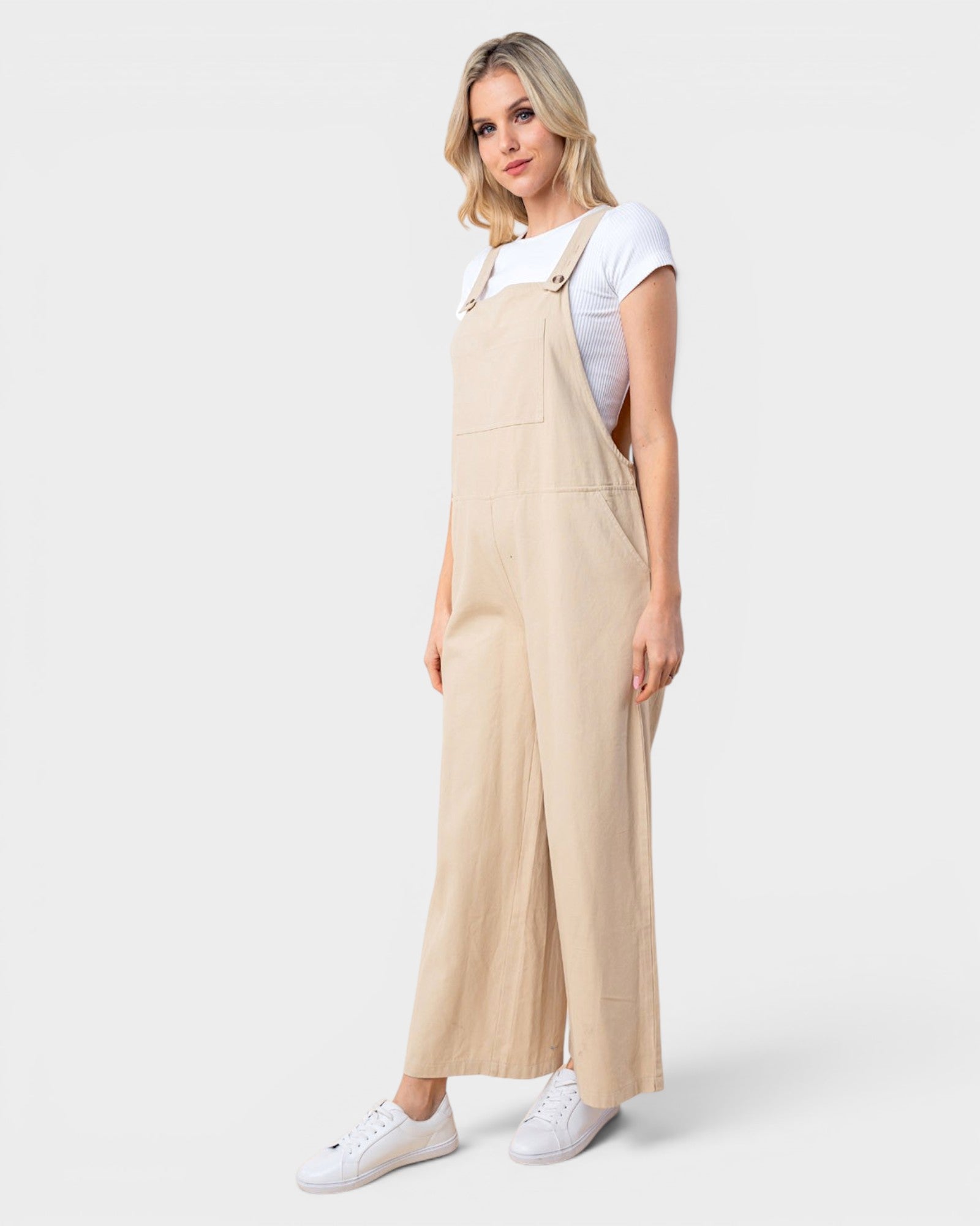 A woman modeling a pair of light beige wide-leg overall jumpsuits over a white t-shirt and white sneakers, photographed against a solid light gray background.