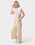 A woman modeling a pair of wide-leg beige linen overall jumpsuit over a white t-shirt and white sneakers, photographed against a solid light gray background.