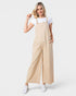A woman modeling a pair of wide-leg beige jumpsuit overalls over a white t-shirt and white sneakers, set against a plain light gray studio background.