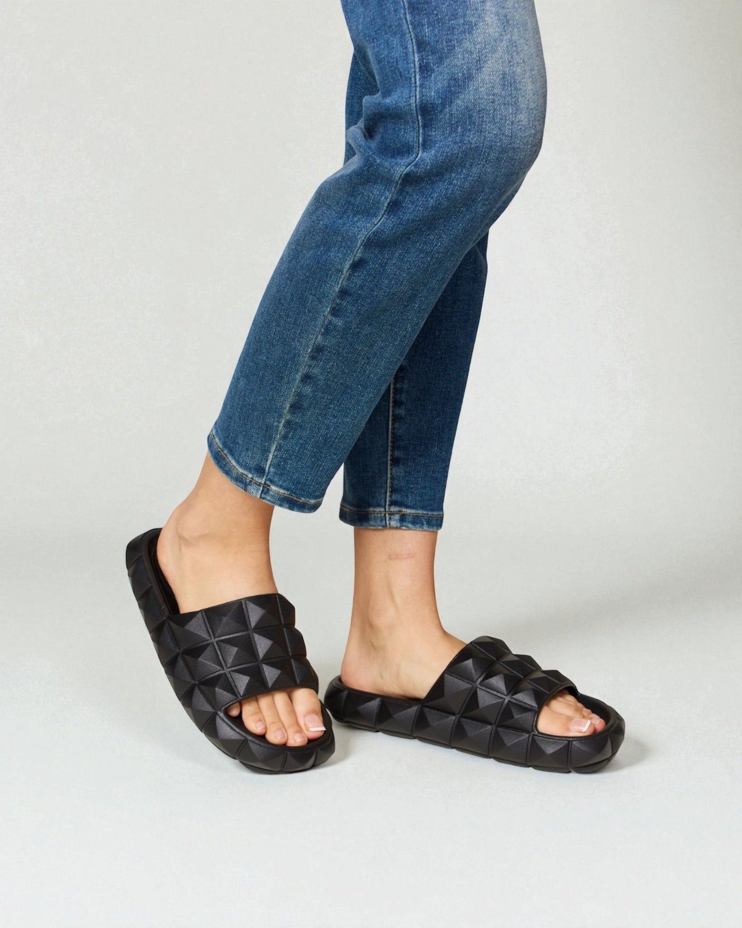 Person wearing black quilted sandals with rolled-up blue jeans on a light gray background