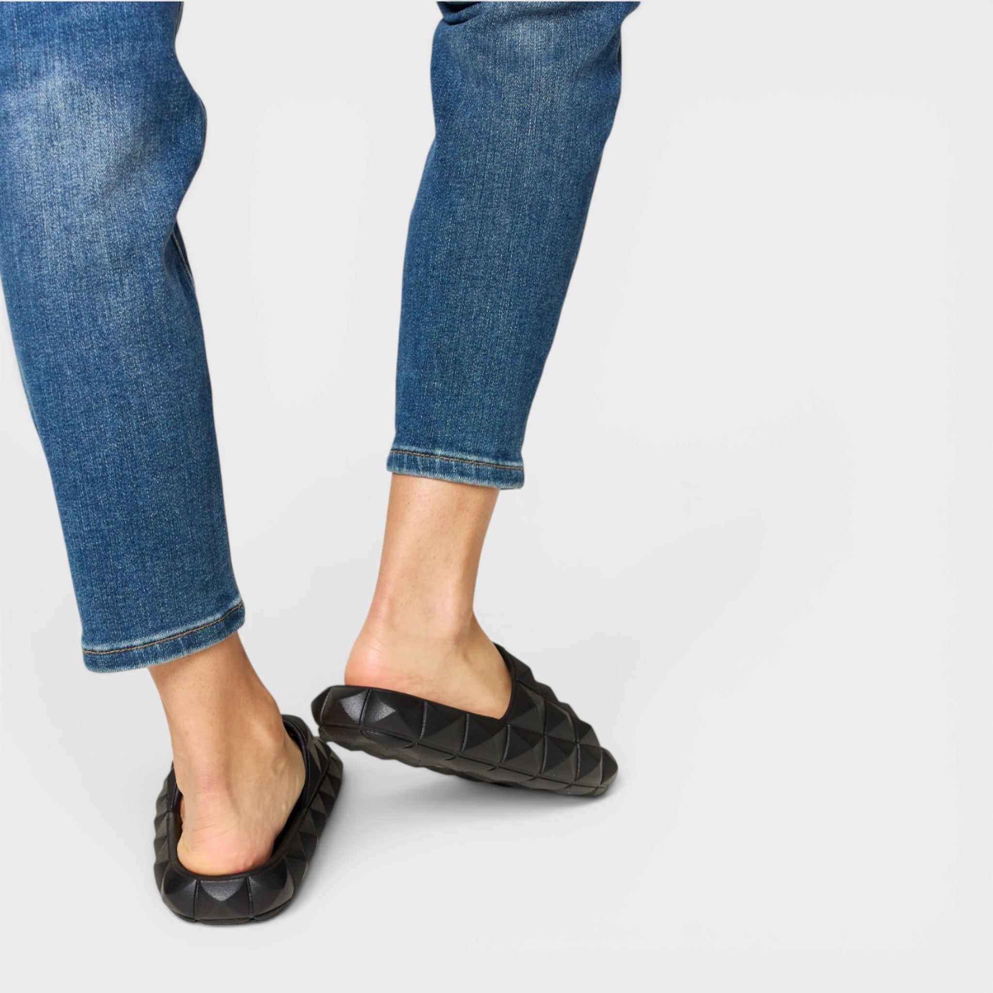 Person wearing black quilted slippers with blue jeans on a light gray background