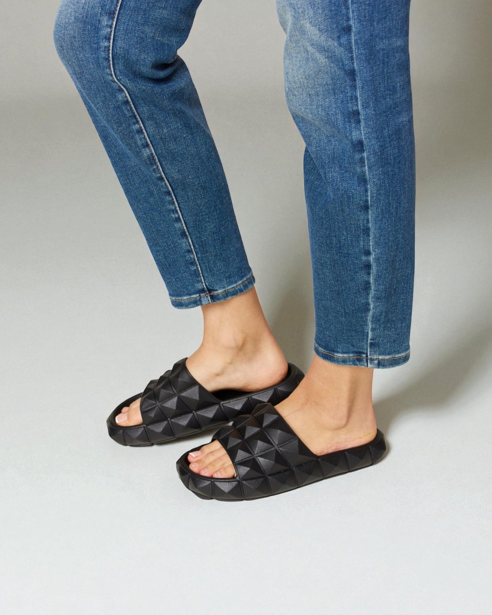 Person wearing black quilted sandals and blue jeans on a light gray background