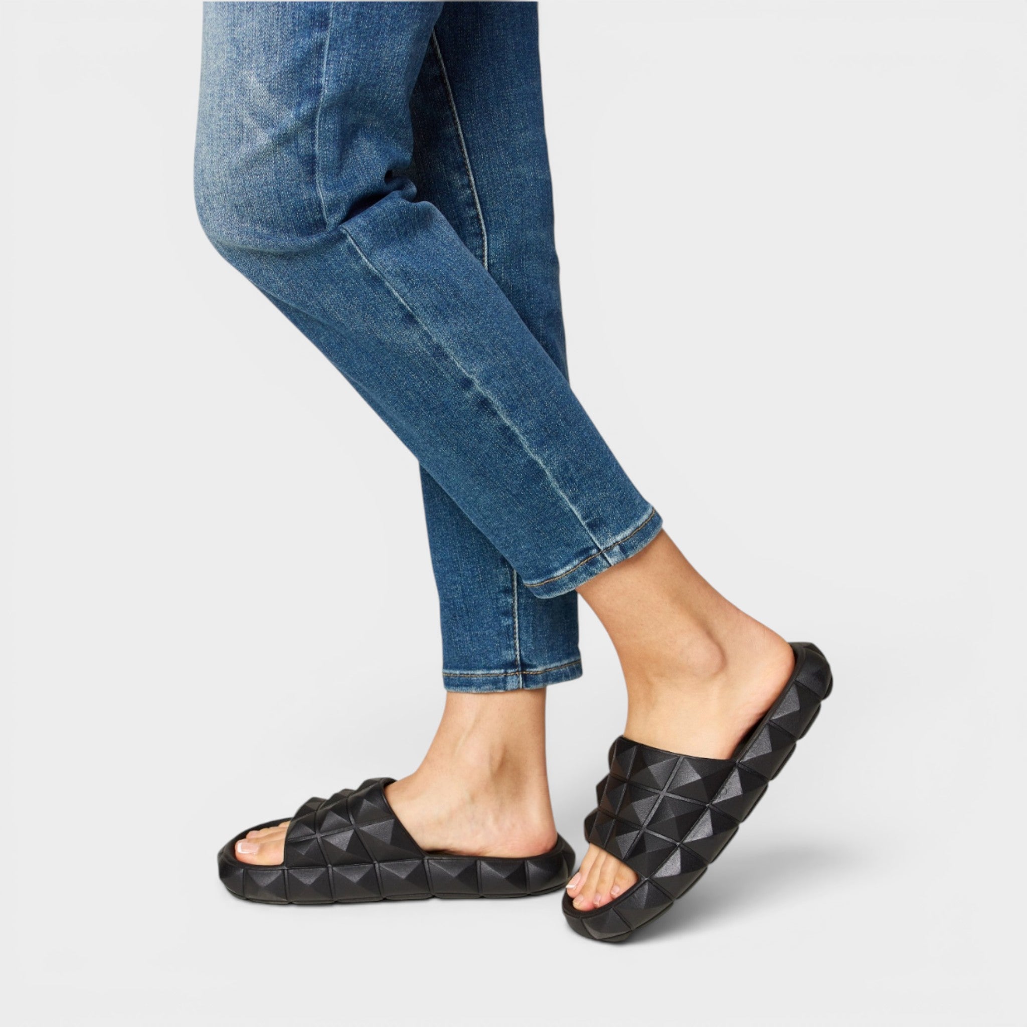 Person wearing black diamond-patterned slides with blue jeans on a light gray background