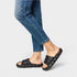 Person wearing black diamond-patterned slides with blue jeans on a light gray background
