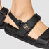 Black sandal with a gold buckle on a light gray background