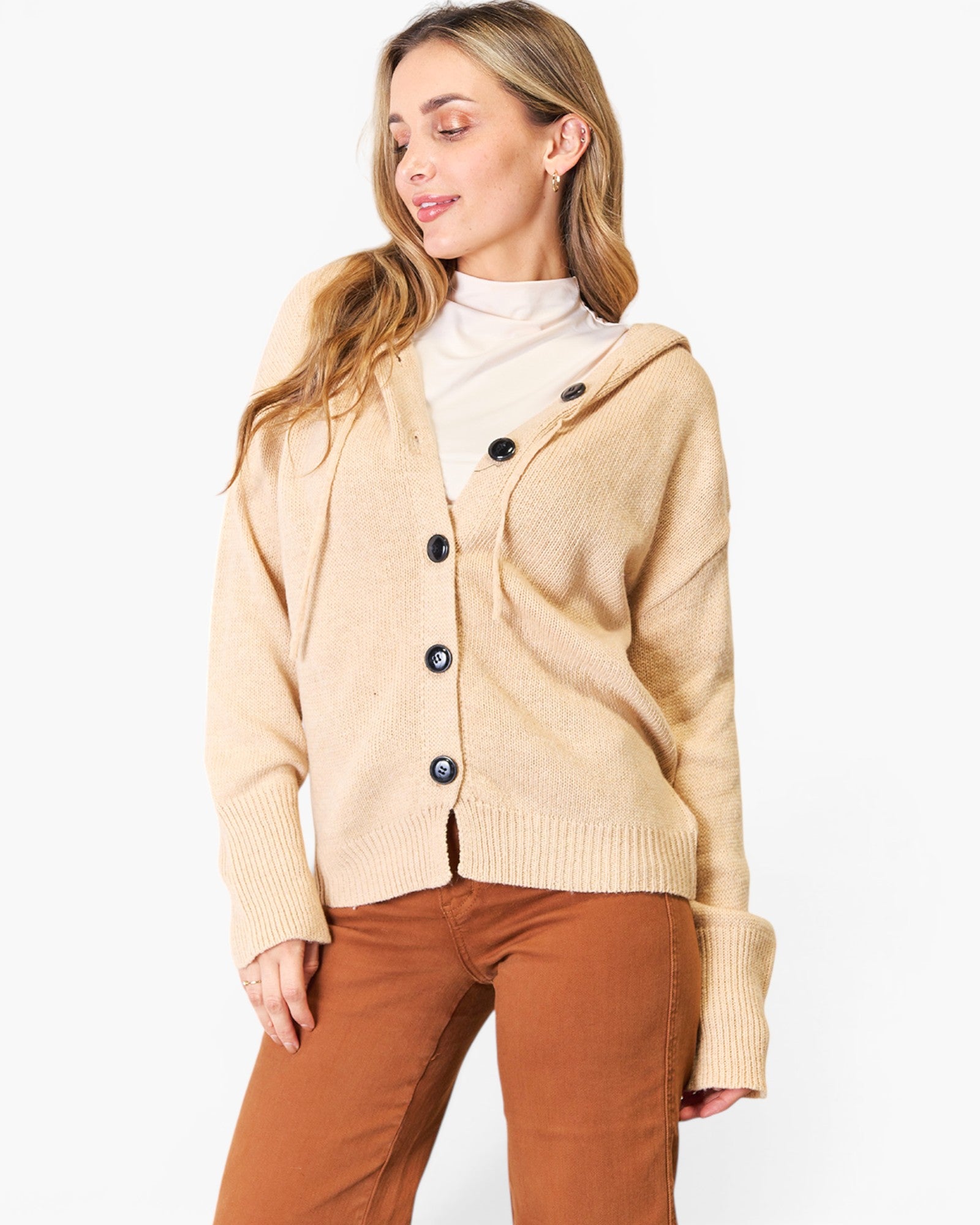 A woman wearing a beige knit cardigan with a hood and dark buttons over a cream turtleneck and rust-brown pants, photographed against a plain white studio background.