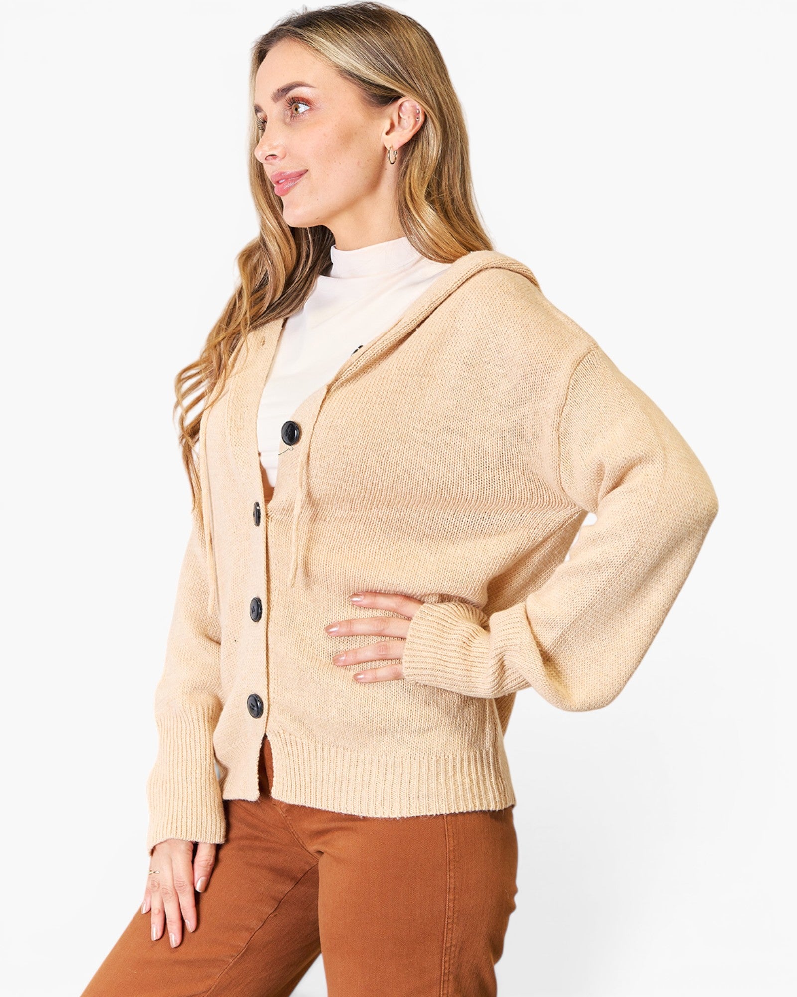 A woman wearing a beige knit cardigan with a hood and front buttons, layered over a white turtleneck and paired with brown pants, photographed against a clean white studio background.