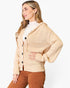 A woman wearing a beige knit cardigan with a hood and front buttons, layered over a white turtleneck and paired with brown pants, photographed against a clean white studio background.