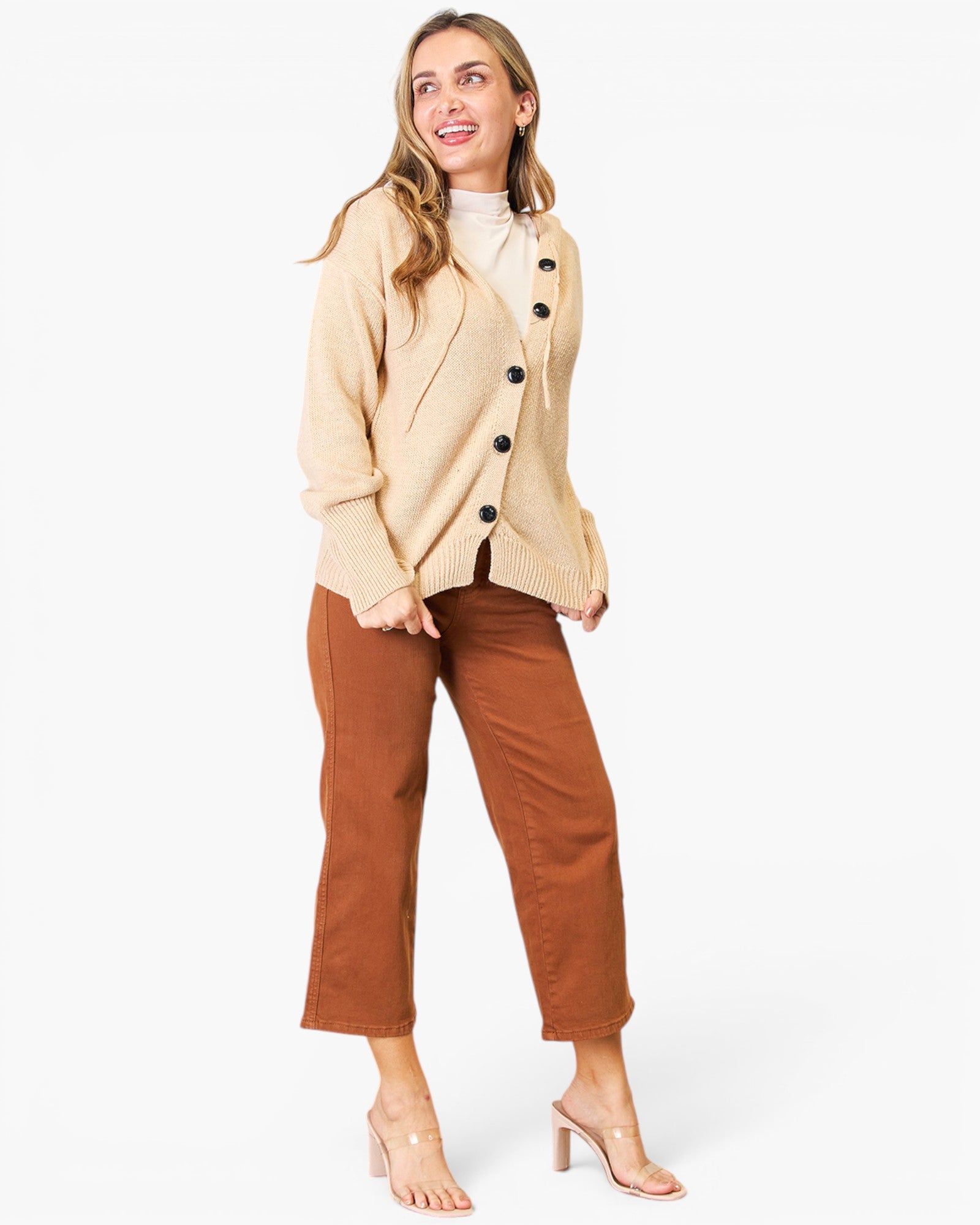 A woman modeling a beige knit cardigan over a white mock neck top, paired with rust-brown cropped pants and clear high-heeled sandals, against a plain white studio background.