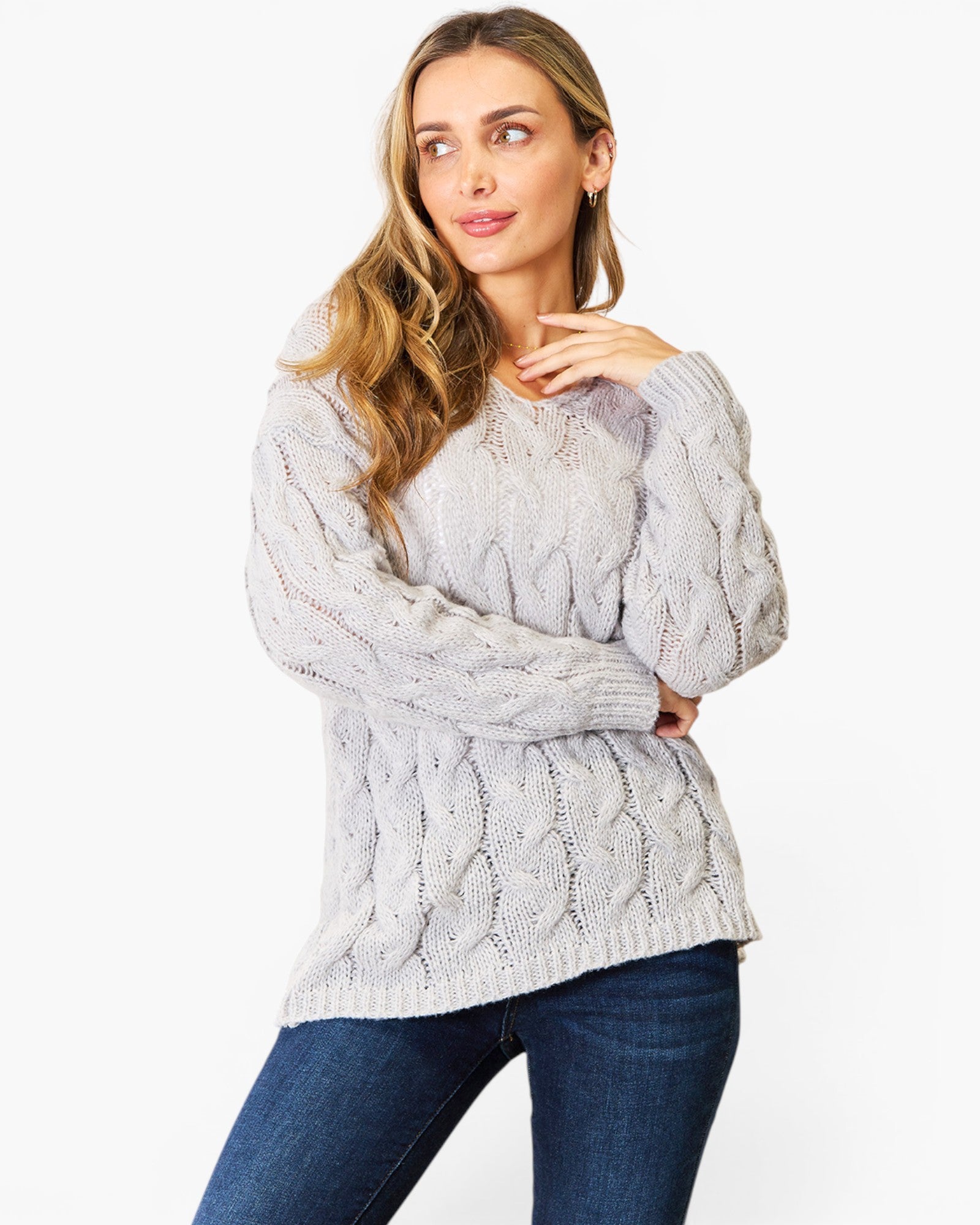 A woman wearing a light gray cable-knit sweater and dark blue jeans, posing against a plain white studio background.