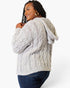 A woman wearing a chunky cable-knit hooded sweater in a light oatmeal gray color, paired with dark blue jeans, shown from a three-quarters back view against a plain white studio background.