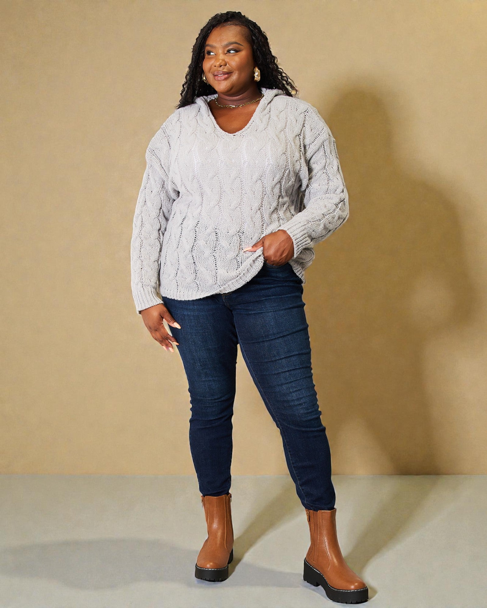 Woman wearing a gray cable knit sweater, blue jeans, and brown boots against a beige background
