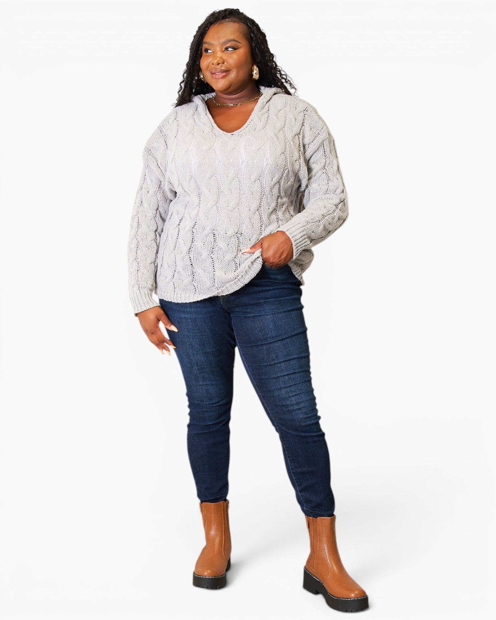 A woman modeling a light gray cable-knit sweater, dark blue skinny jeans, and brown ankle boots against a plain white studio background.
