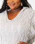 A close-up of a woman wearing a light gray cable-knit sweater with a V-neck, styled with gold jewelry against a plain white background.