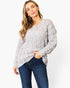 A woman wearing a light gray cable-knit sweater and dark blue jeans, standing against a plain white studio background.