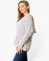 A woman wearing a light gray cable-knit sweater and dark blue jeans, photographed against a plain white studio background.