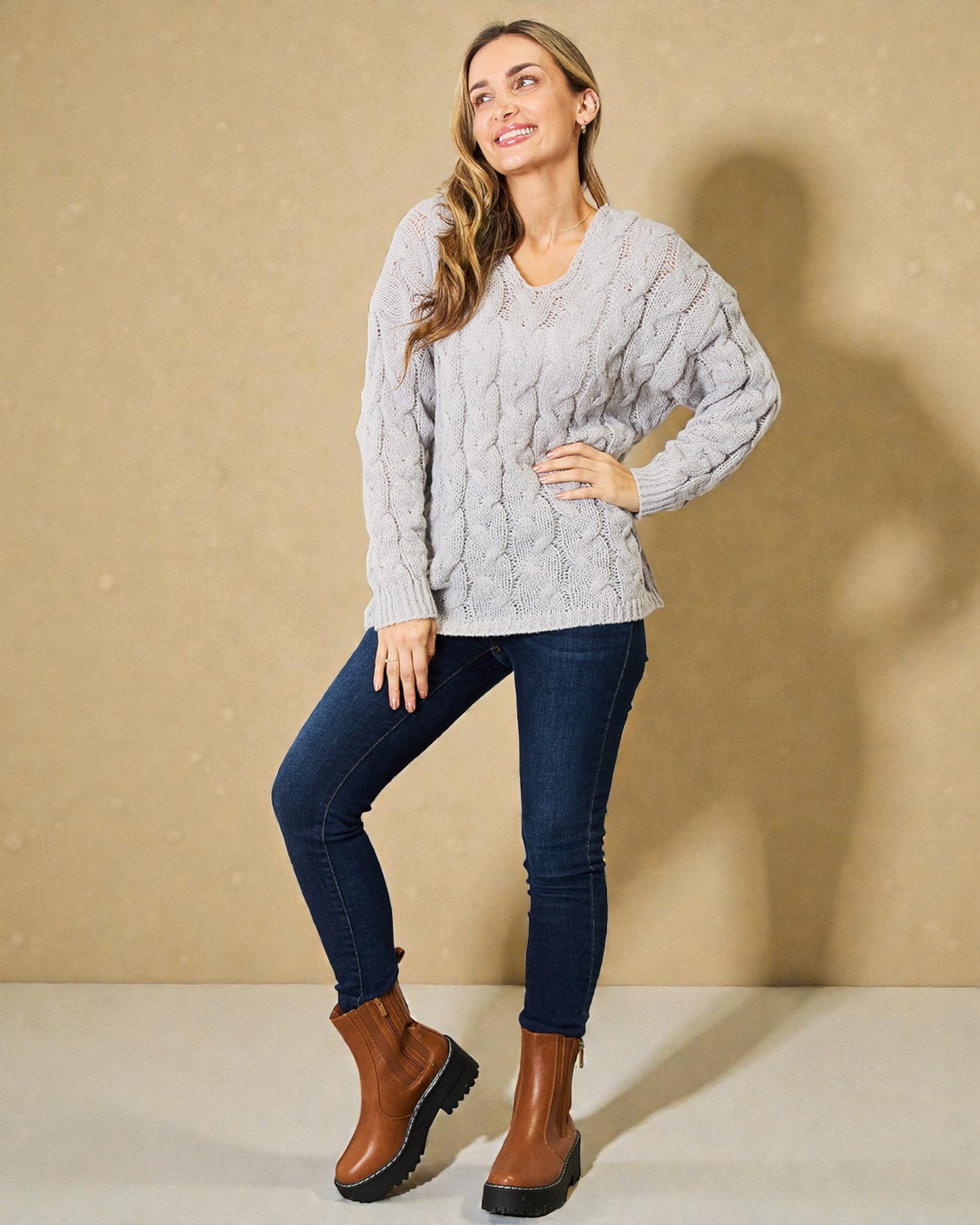 Woman wearing a gray cable knit sweater, blue jeans, and brown boots against a beige background