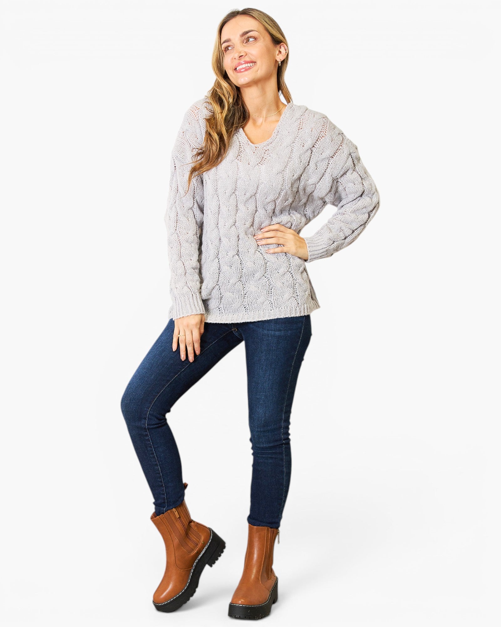A woman modeling a light gray cable-knit sweater, dark blue skinny jeans, and brown ankle boots against a plain white studio background.