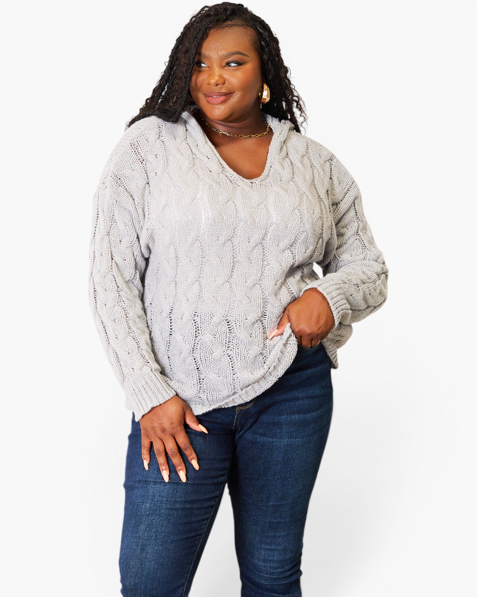 A woman modeling a light gray cable-knit sweater with a V-neck and long sleeves, paired with dark blue jeans, against a plain white background.