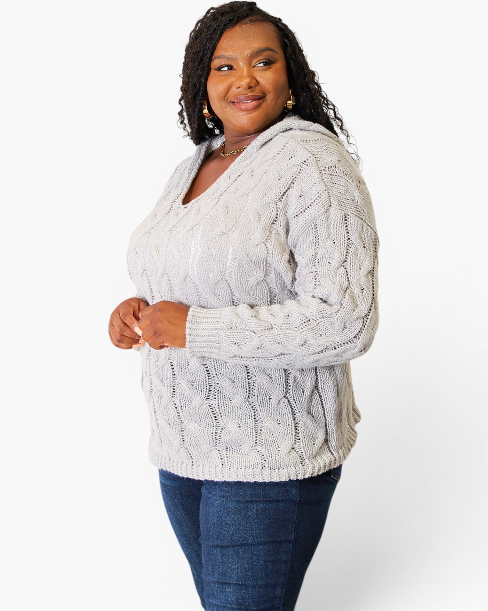 A woman modeling a light gray cable-knit hooded sweater with blue jeans, photographed against a clean white studio background.