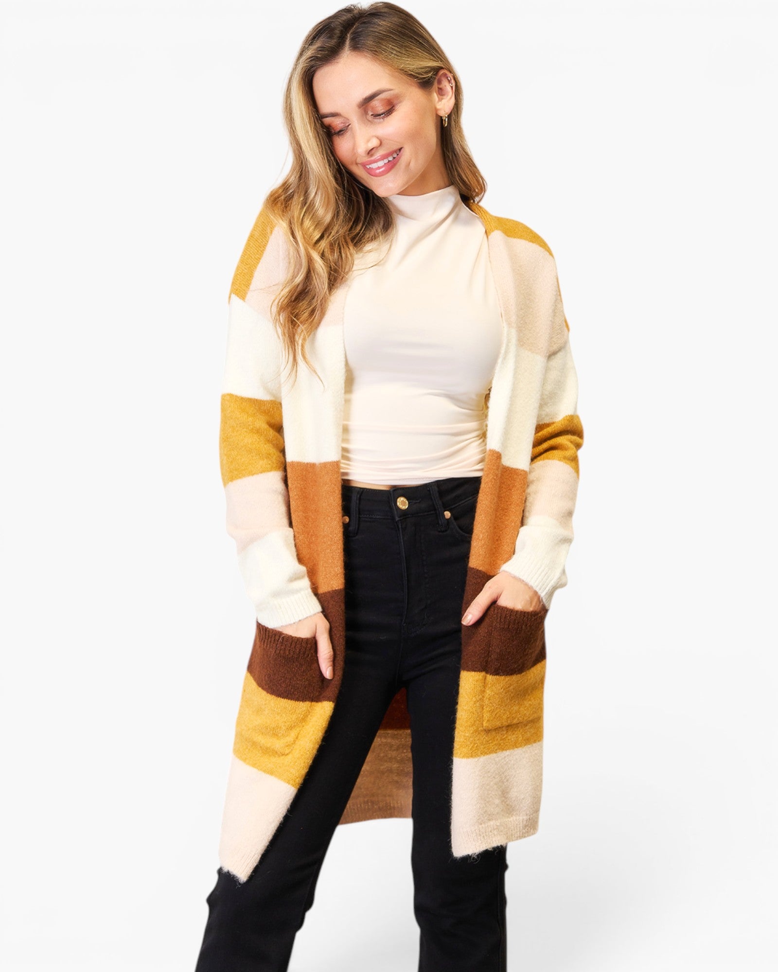 A woman wearing a long open-front knit cardigan with large color-block panels in cream, mustard yellow, tan, and brown, paired with a cream turtleneck and black jeans, against a plain white studio background.
