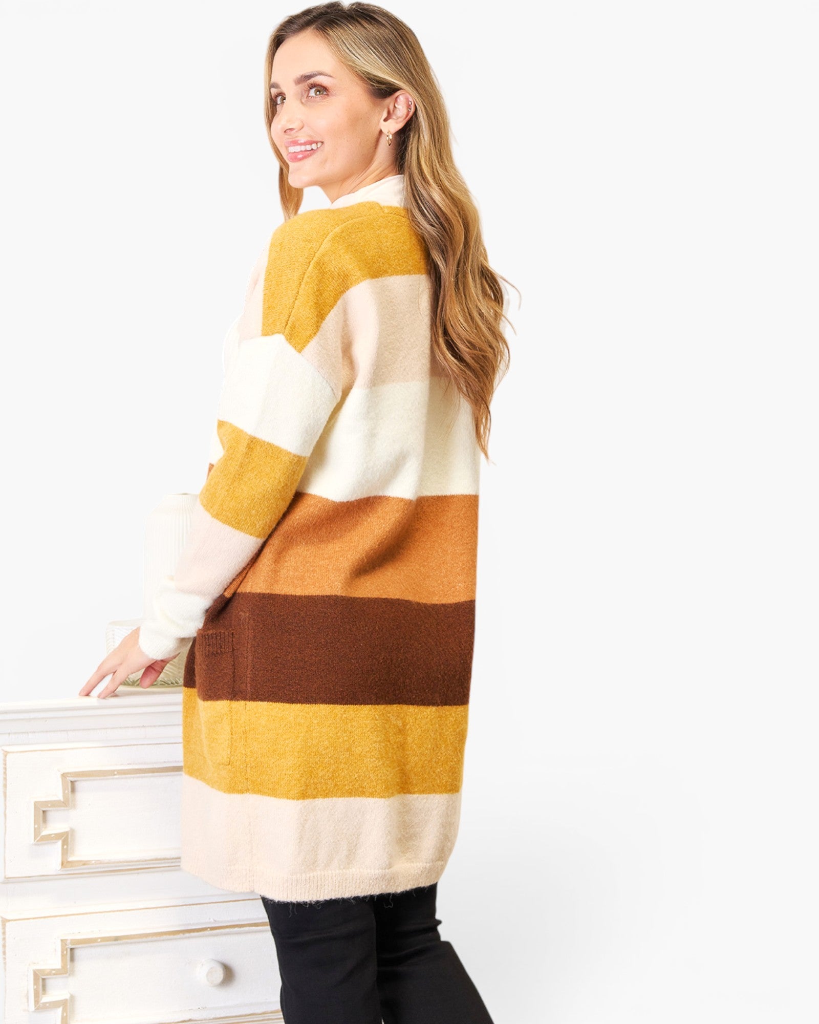 A woman wearing a fuzzy knit sweater with wide horizontal stripes in cream, mustard yellow, orange, and brown, standing next to a white dresser against a white background.