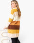 A woman wearing a fuzzy knit sweater with wide horizontal stripes in cream, mustard yellow, orange, and brown, standing next to a white dresser against a white background.