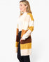 A woman wearing a chunky knit color-block cardigan with stripes of mustard yellow, cream, and brown over a white top and black pants, photographed against a plain white studio background.