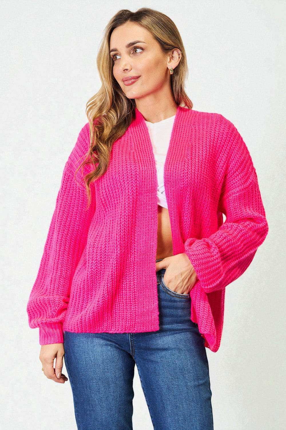 Woman wearing a bright pink cardigan over a white top and blue jeans on a light gray background