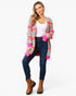A woman modeling a gray striped cardigan with bright pink and orange accents, worn over a white tank top and dark blue jeans, paired with brown ankle boots, on a light gray background.