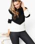 A woman wearing a black and white color-block knit sweater and black pants, leaning against a white table in a bright indoor setting.