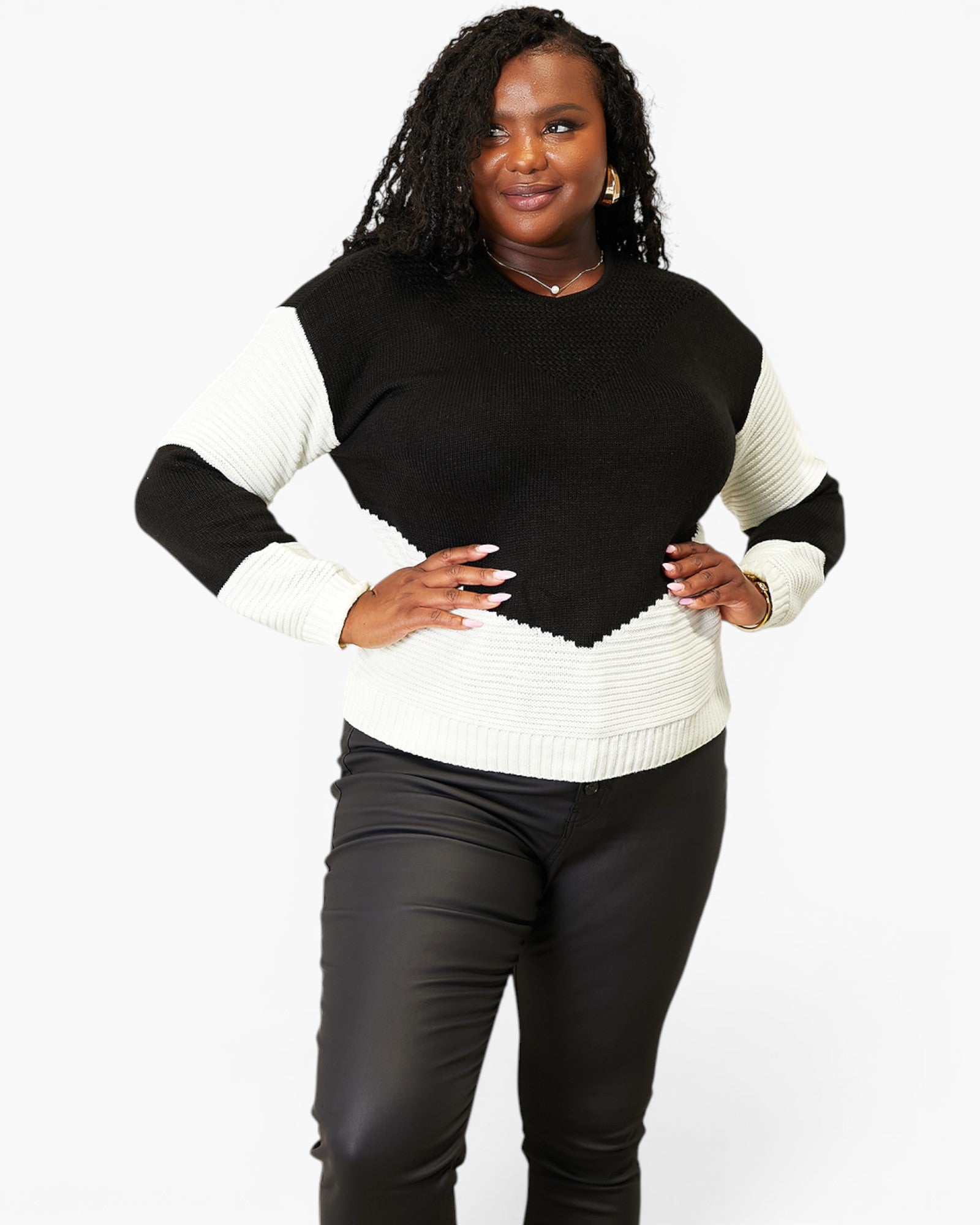 A woman modeling a color-block knit sweater with a black yoke and black ribbed sleeves, paired with black pants, against a plain white studio background.