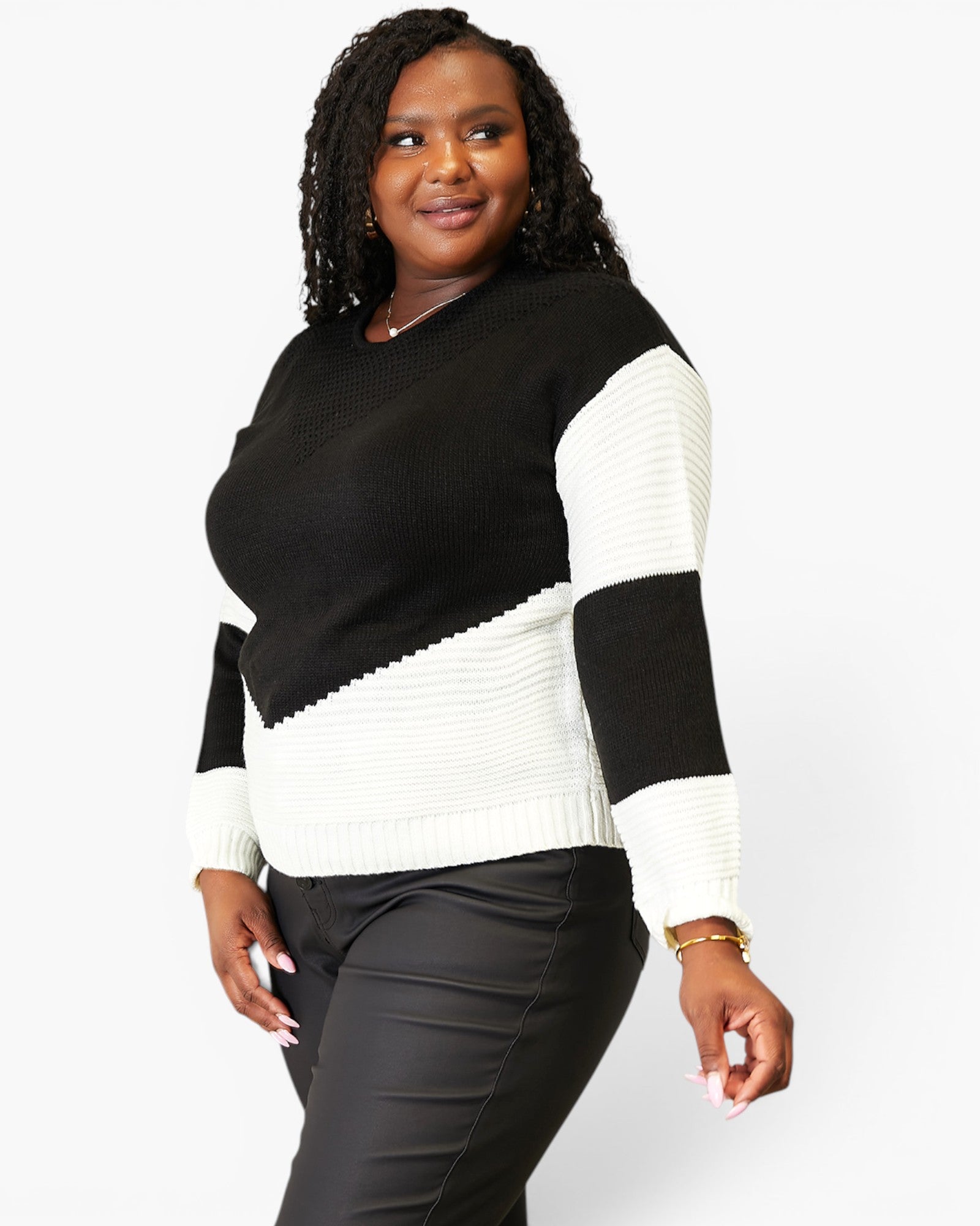 A woman modeling a color-block knit sweater with a black body, black shoulder panel, and white ribbed sleeves and lower body, paired with black pants against a light gray studio background.