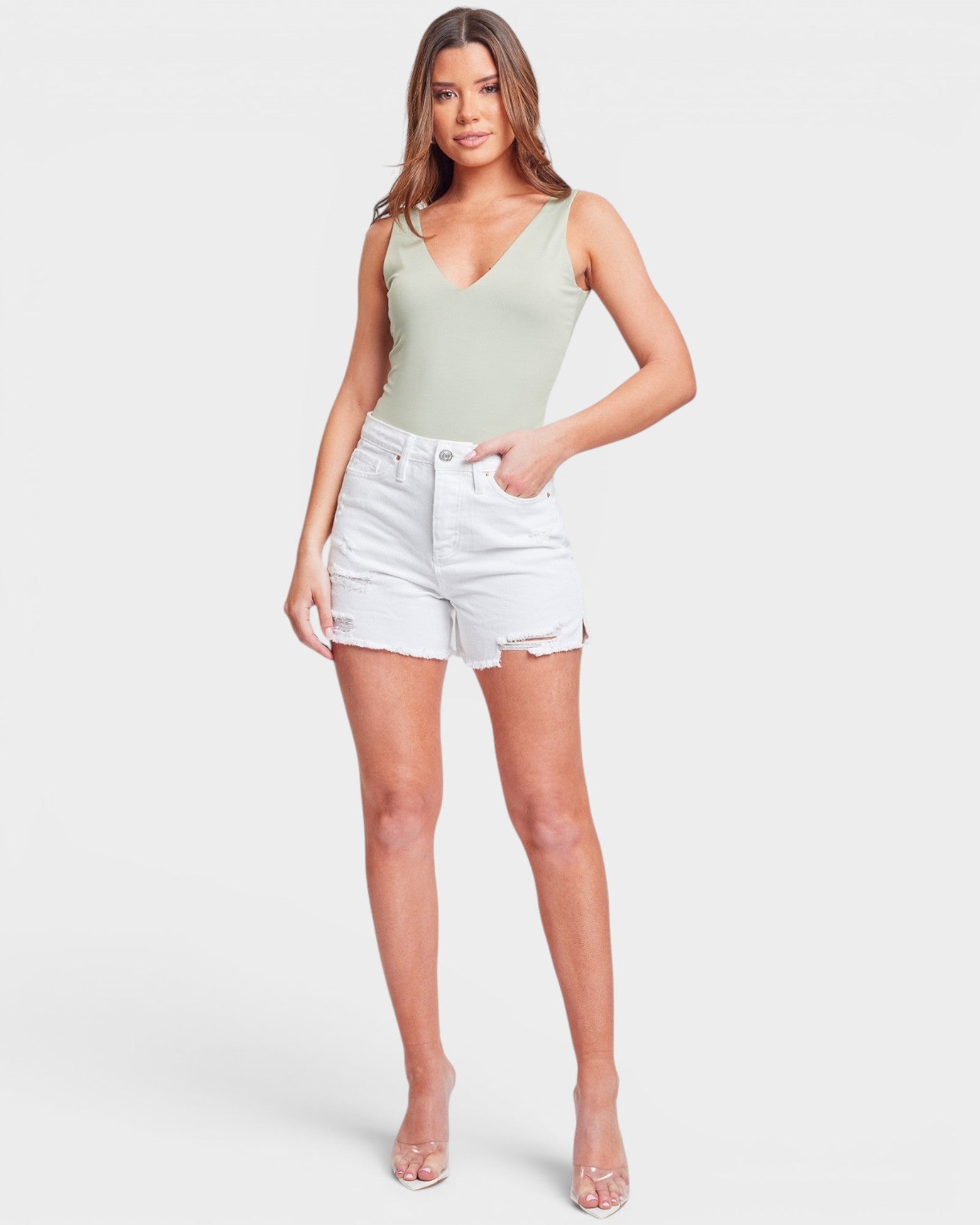A woman modeling a light sage green ribbed tank top and white denim shorts against a light gray studio background.