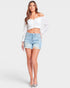 A woman modeling a white off-the-shoulder cropped long-sleeve top and light-wash distressed denim shorts against a light gray studio background.