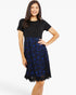 A woman modeling a short-sleeve knee-length dress with a solid black bodice and a blue and black floral lace skirt, set against a plain white studio background.