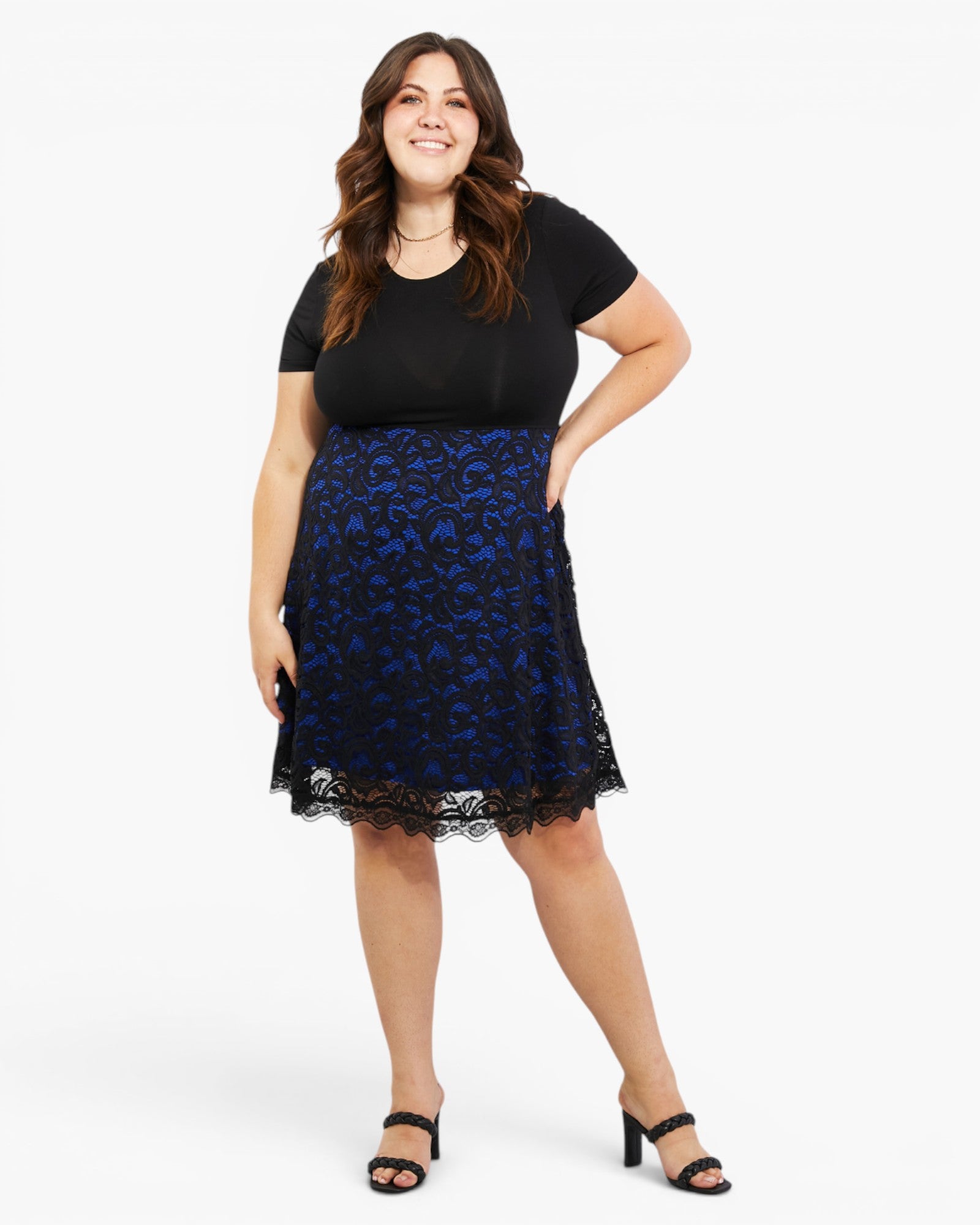 A woman modeling a black short-sleeve top and a blue floral lace skirt with black lace trim, paired with black heeled sandals, against a plain white studio background.