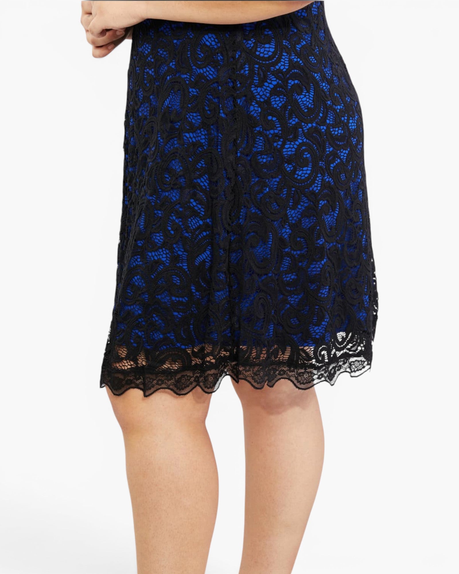 A close-up of a model wearing a knee-length black lace skirt with a vibrant blue floral lace overlay and scalloped black lace hem, set against a plain white background.